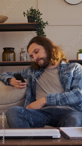 Bored man using a cellphone for online leisure and browsing the internet at home, seeking entertainment to alleviate monotony in a familiar setting. Vertical video.