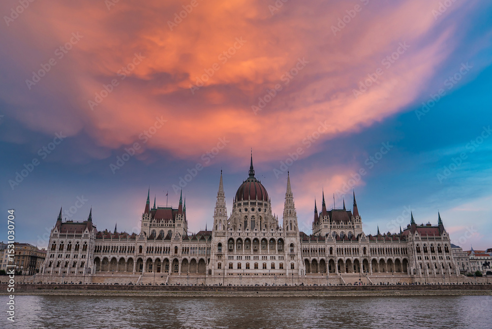 Fototapeta premium The neo Gothic Hungarian Parliament Building in Budapest, Hungary, stands by the Danube River under a vibrant orange and pink sky with calm reflections.