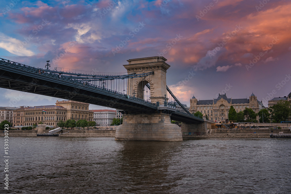 Fototapeta premium The Szechenyi Chain Bridge spans the Danube River in Budapest, Hungary, with stone towers, suspension cables, and a vibrant sunset sky reflected in the water.