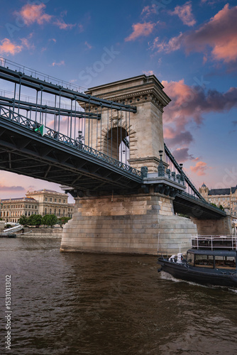 The Szechenyi Chain Bridge spans the Danube River in Budapest, Hungary, with stone towers, suspension cables, and a vibrant sunset sky. A boat is on the river.