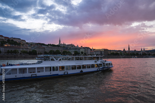A river cruise boat glides along the Danube River at sunset in Budapest, with Matthias Church and historic buildings visible on the Buda side.