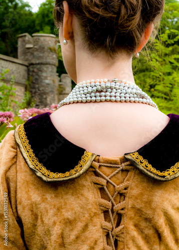 A woman in a historical costume, wearing a pearl necklace and earrings, stands with her back to the camera.