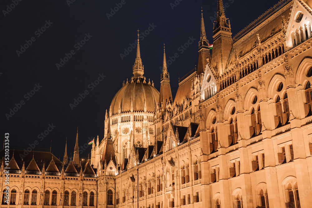 Fototapeta premium The Hungarian Parliament Building in Budapest glows at night, showcasing neo Gothic architecture with spires, a central dome, and intricate details.