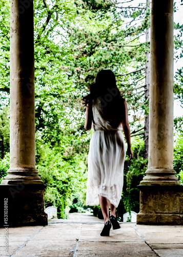 A woman in a white dress walks away from the camera through a stone archway.