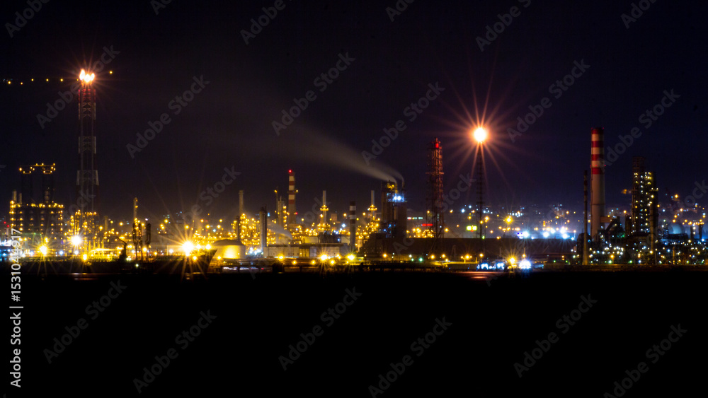 Fototapeta premium Night view of an illuminated oil refinery with glowing chimneys, industrial buildings, and flaring towers under a dark sky, showcasing the energy and manufacturing sector's nighttime operations.