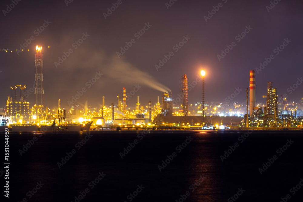 Naklejka premium Brightly lit oil refinery at night with smoke emissions, flaring towers, and glowing chimneys against a dark sky, illustrating industrial activity and energy production in full operation.
