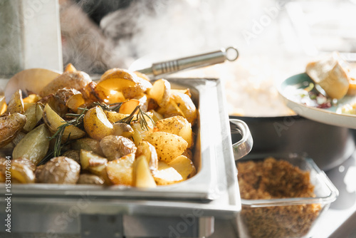 Close-up of roasted potatoes with rosemary in a buffet tray, with steam rising in a warm catering setting.
