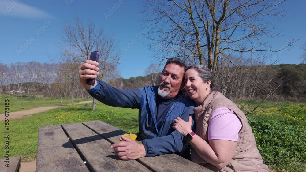 Happy mature couple doing a videocall on a picnic table in the park Happy mature couple doing a videocall on a picnic table in the park