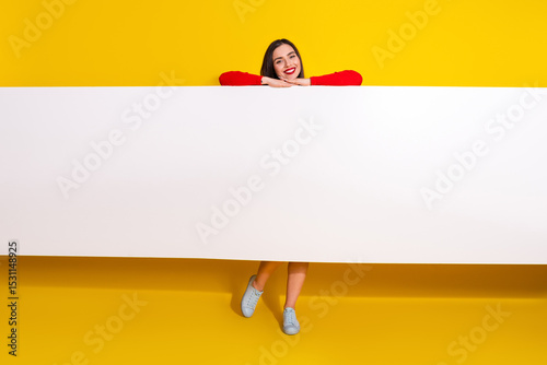 Young woman posing with a blank sign on a vibrant yellow background, wearing a red shirt and sneakers, expressing cheerfulness and charm.