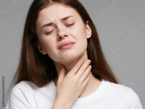 Close-up of a young woman with long brown hair, wearing a white t-shirt, holding her throat in pain against a gray background, concept for health and medical advertisement