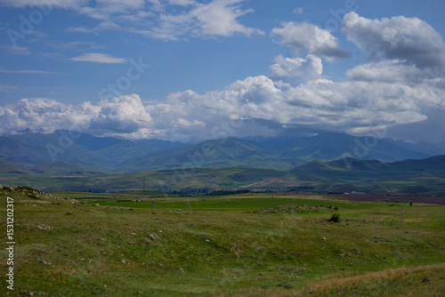 Scenic view of lush green hills and dramatic red cliffs under a bright blue sky with white clouds, showcasing the beauty of untouched mountainous nature and diverse rocky terrain.