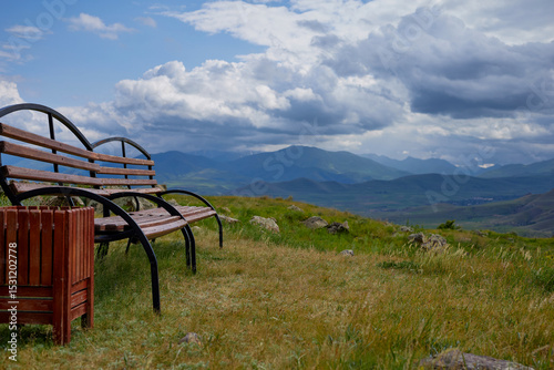Scenic mountain view with two empty wooden benches and a trash bin on a grassy hilltop under dramatic clouds, offering a peaceful and serene natural landscape perfect for relaxation and reflection.