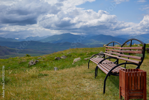 Scenic mountain view with two empty wooden benches and a trash bin on a grassy hilltop under dramatic clouds, offering a peaceful and serene natural landscape perfect for relaxation and reflection.