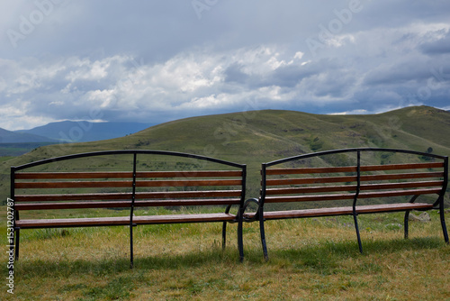 Scenic mountain view with two empty wooden benches and a trash bin on a grassy hilltop under dramatic clouds, offering a peaceful and serene natural landscape perfect for relaxation and reflection.