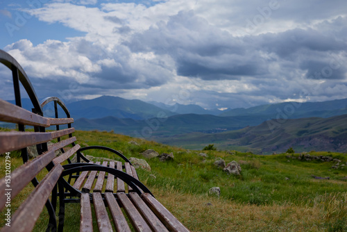Scenic mountain view with two empty wooden benches and a trash bin on a grassy hilltop under dramatic clouds, offering a peaceful and serene natural landscape perfect for relaxation and reflection.