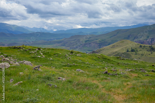 Scenic view of lush green hills and dramatic red cliffs under a bright blue sky with white clouds, showcasing the beauty of untouched mountainous nature and diverse rocky terrain.