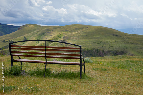Scenic mountain view with two empty wooden benches and a trash bin on a grassy hilltop under dramatic clouds, offering a peaceful and serene natural landscape perfect for relaxation and reflection.