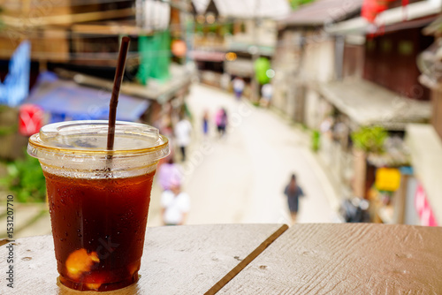 Fototapeta Naklejka Na Ścianę i Meble -  Iced coconut americano coffee on wooden table with blurry Mae Kampong street background, small village with traditional houses and beautiful nature. Destination of traveler. Chiang Mai, Thailand.