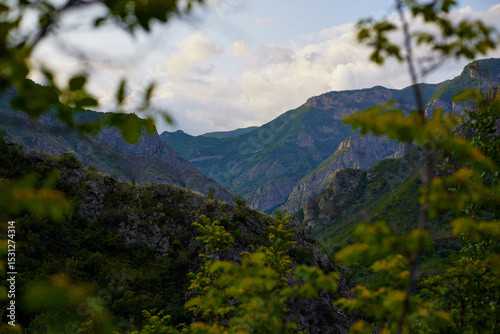 Scenic view of lush green hills and dramatic red cliffs under a bright blue sky with white clouds, showcasing the beauty of untouched mountainous nature and diverse rocky terrain.
