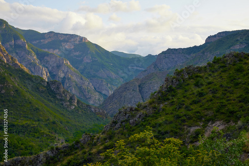 Scenic view of lush green hills and dramatic red cliffs under a bright blue sky with white clouds, showcasing the beauty of untouched mountainous nature and diverse rocky terrain.