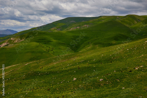 Scenic view of lush green hills and dramatic red cliffs under a bright blue sky with white clouds, showcasing the beauty of untouched mountainous nature and diverse rocky terrain.