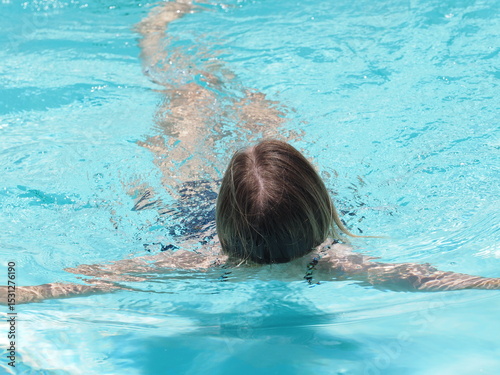 Woman relaxing in outside swimming pool during hot daytime. Back view.