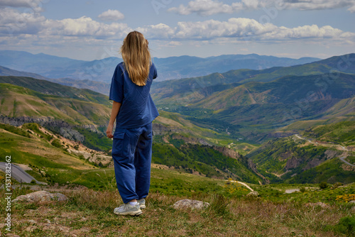 Woman standing on a mountain peak with arms outstretched, enjoying a breathtaking panoramic view of green hills and valleys under a partly cloudy sky, symbolizing freedom and connection with nature.
