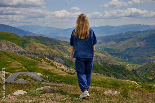 Woman standing on a mountain peak with arms outstretched, enjoying a breathtaking panoramic view of green hills and valleys under a partly cloudy sky, symbolizing freedom and connection with nature.