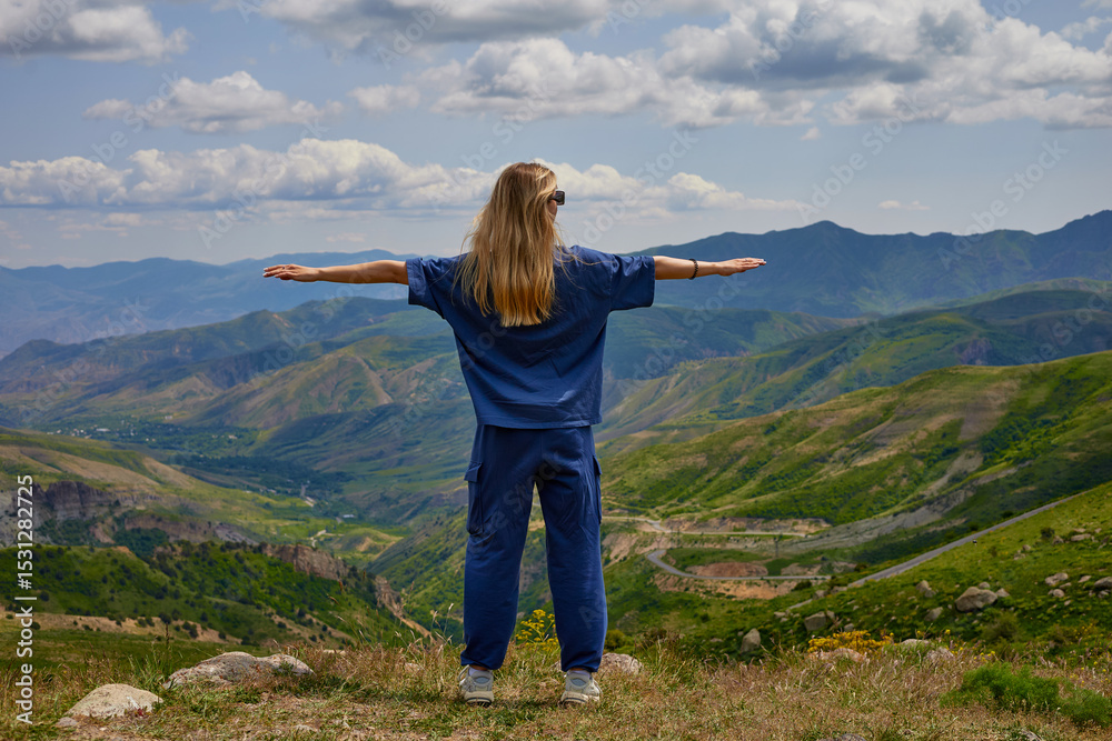 Naklejka premium Woman standing on a mountain peak with arms outstretched, enjoying a breathtaking panoramic view of green hills and valleys under a partly cloudy sky, symbolizing freedom and connection with nature.