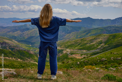 Woman standing on a mountain peak with arms outstretched, enjoying a breathtaking panoramic view of green hills and valleys under a partly cloudy sky, symbolizing freedom and connection with nature.