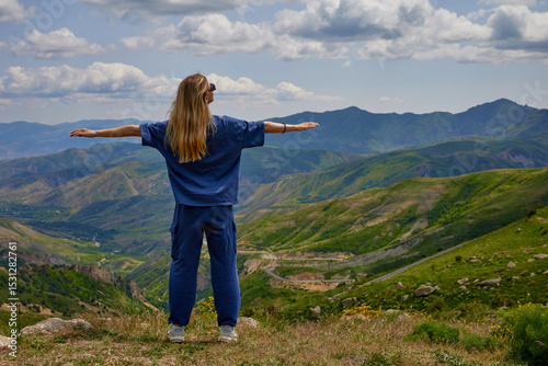 Woman standing on a mountain peak with arms outstretched, enjoying a breathtaking panoramic view of green hills and valleys under a partly cloudy sky, symbolizing freedom and connection with nature.