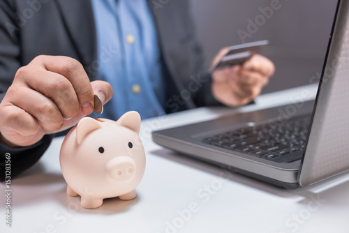 A person inserts a coin into a piggy bank while holding a credit card near a laptop on a desk.