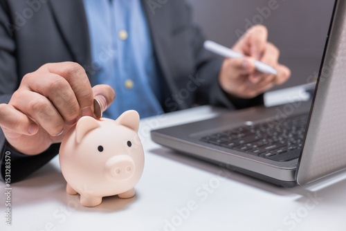 A person in business attire inserts a coin into a piggy bank beside a laptop, symbolizing saving money and financial management.