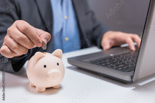 A person in business attire places a coin into a piggy bank beside a laptop, symbolizing saving money and financial management.