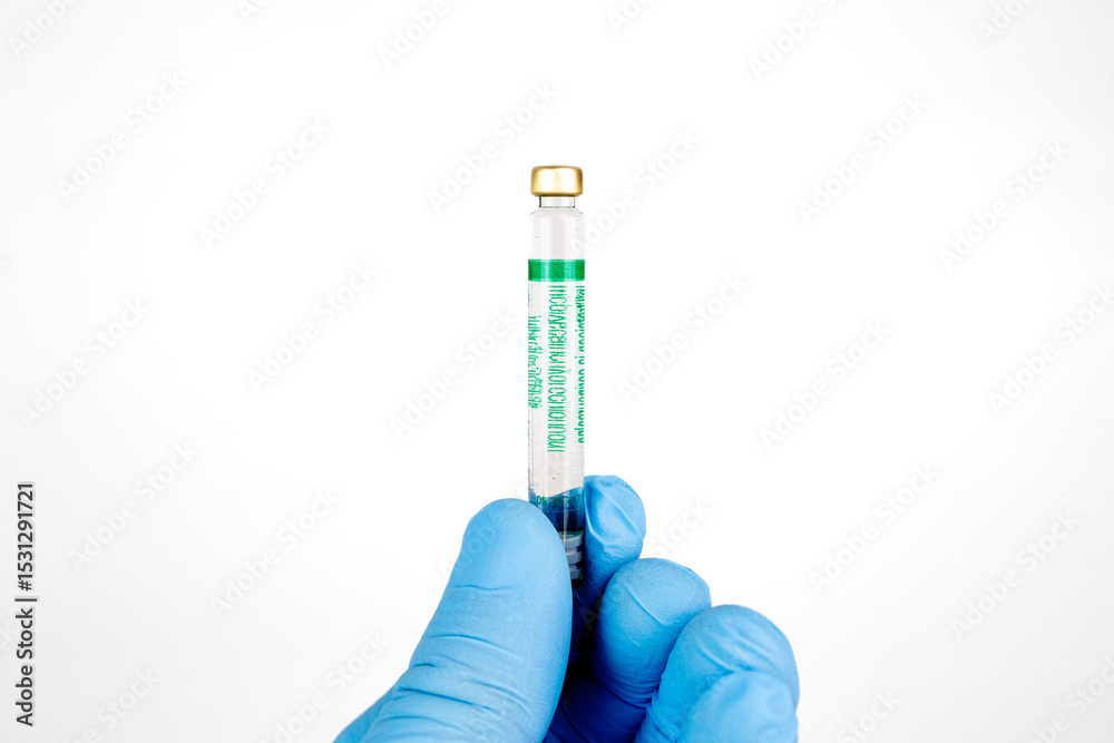 Obraz premium Gloved Hand Holding Medical Vaccine Vial on White Background, Dental Anesthesia Syringe.Close-up of a blue-gloved hand holding a small glass vaccine vial with a green label