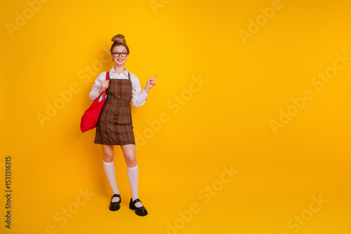 Cheerful young student wearing stylish school uniform standing confidently with a backpack against yellow background pointing excitedly