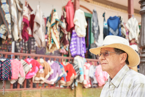 Portrait of a senior Hispanic man, his expression is one of concern. He is outdoors and behind him is an artisan clothing store displaying ruanas, ponchos and sweaters of various colors and styles.