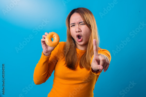 Asian woman in orange sweater holding a glazed donut in one hand and raising her index finger with a serious expression, against a bright blue background