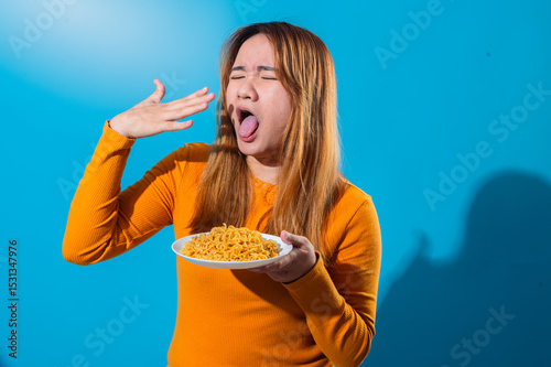 Asian woman in orange shirt with a disgusted expression, sticking out tongue while holding a plate of instant noodles, reacting negatively to the food against a blue background