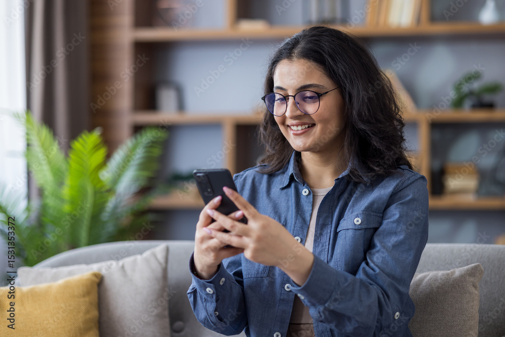 Smiling young Indian woman sitting on sofa at home and using mobile phone Smiling young Indian woman sitting on sofa at home and using mobile phone