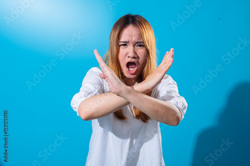 Asian woman in a white shirt with serious expression crossing her arms in front of her chest to form an X gesture, signaling disapproval or prohibition, against a solid blue background