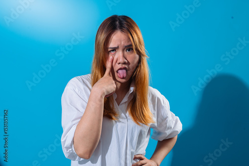 Asian woman in a white shirt pulling her lower eyelid down with one finger and sticking out her tongue in a silly and exaggerated expression, standing against a solid blue background