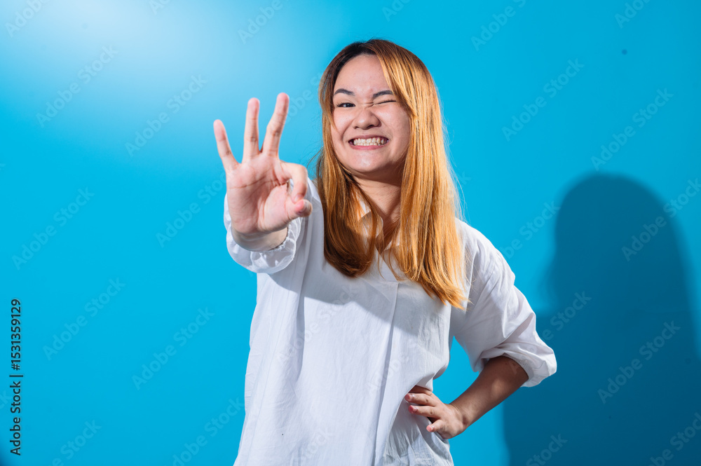 Fototapeta premium Asian woman with long straight hair in a white shirt smiles brightly while making an OK hand gesture with one hand and winking playfully against a plain blue background in studio lighting