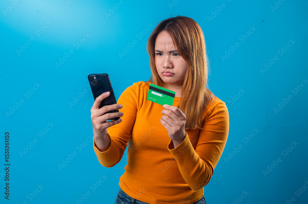 Asian woman in orange shirt looks confused and frustrated while checking her phone and holding a green credit card, against a solid blue background in a studio shot Asian woman in orange shirt looks confused and frustrated while checking her phone and holding a green credit card, against a solid blue background in a studio shot