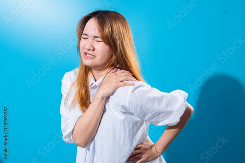 Asian woman wincing in pain while holding her shoulder and back, wearing a white shirt with eyes closed, conveying discomfort or injury, set against a bright blue studio background