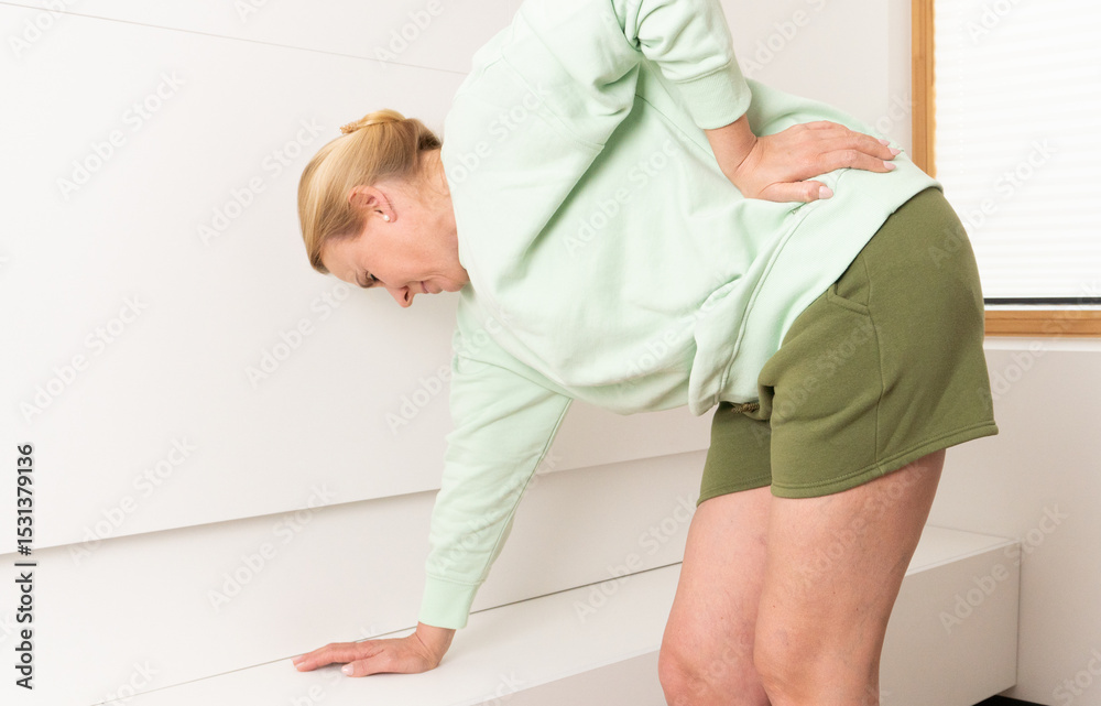 Fototapeta premium Mature woman bending forward and holding her lower back in pain. Concept of spine discomfort, back injury, posture strain or chronic pain in daily life at home.