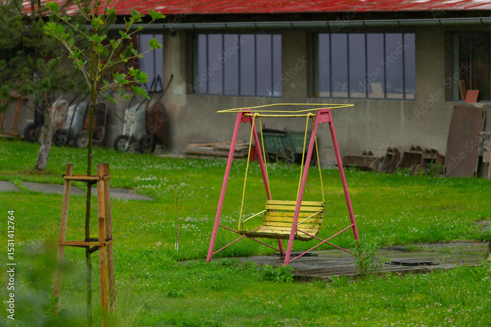 Fototapeta premium In a vibrant green field, a lonely yellow swing set sits among trees and a distant building, symbolizing an unused play space longing for laughter and nostalgic childhood memories