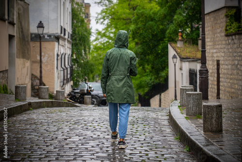 Woman in dark green raincoat and blue pants walking cobblestone streets of Montmartre Paris with spring trees