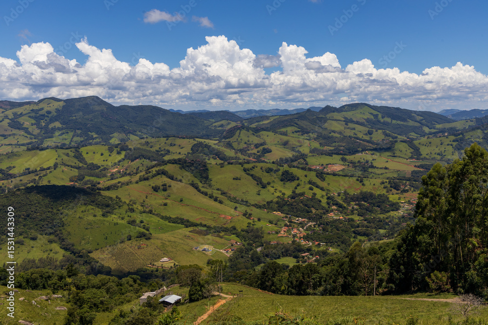 Obraz premium A scenic landscape of rolling green hills and mountains captured in Extrema, Minas Gerais, Brazil. The image shows lush pastureland, forest patches, and dramatic cloud formations against a deep blue