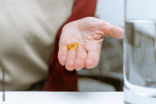 Macro shot of woman's hand holding omega-3 softgel capsule. Focus is entirely on transparent capsule resting on her palm, glass of water softly blurred in foreground Daily health routines, self-care